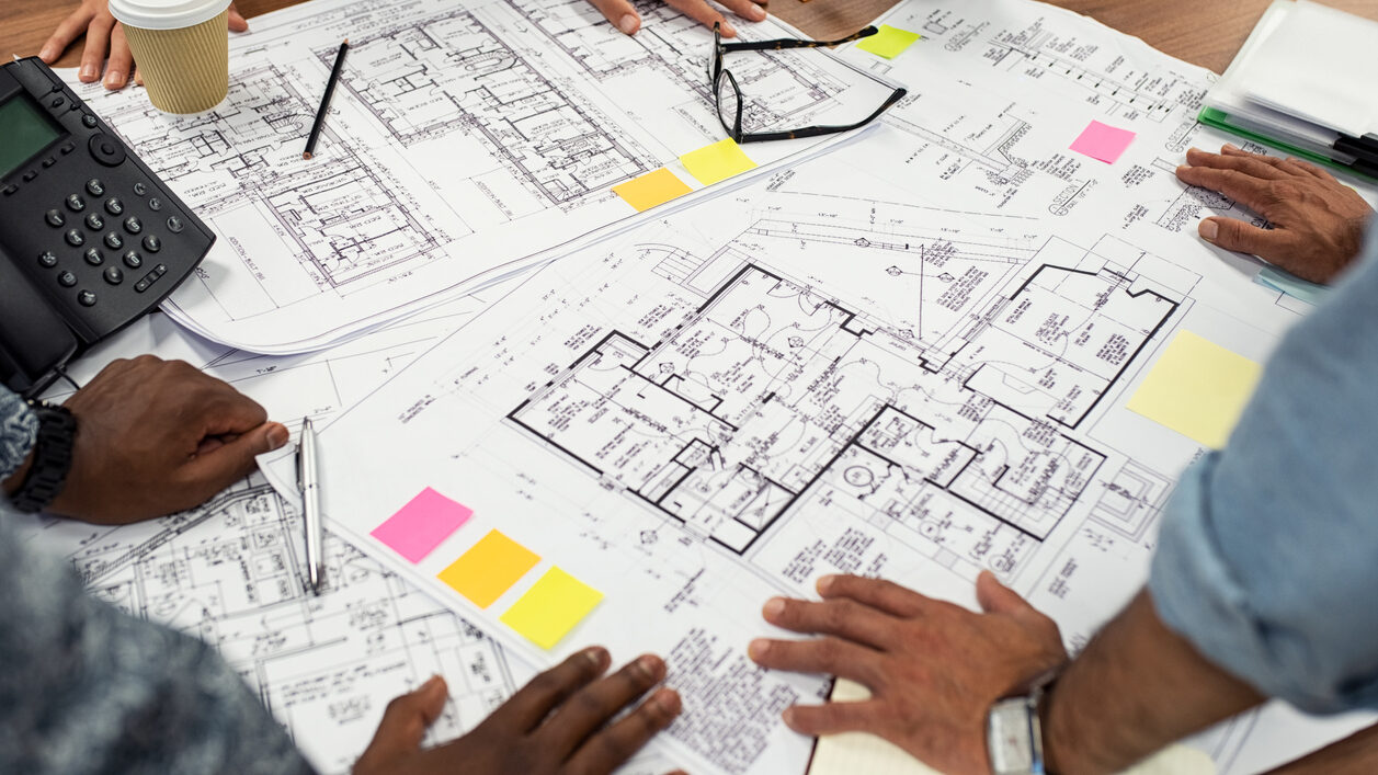 Three people are analyzing house plans on a table. You cannot see their faces, just their hands on the table where the plans are located.