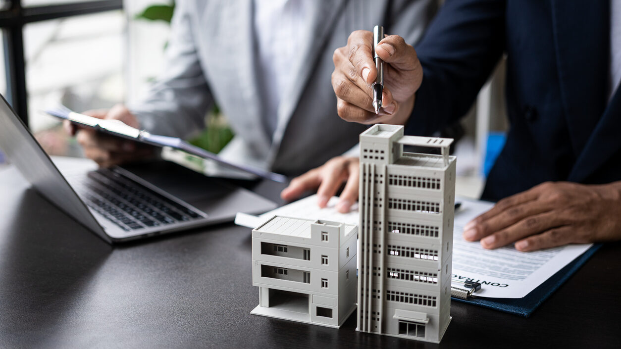 A real estate agent points to a building model located on a table with a pencil while another agent is sitting behind him in front of her laptop.